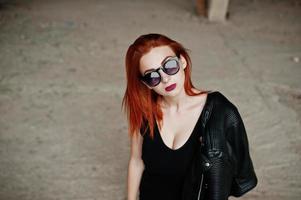 chica elegante de pelo rojo con gafas de sol vestidas de negro, contra un lugar abandonado. foto