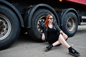 Chica elegante de pelo rojo vestida de negro, sentada contra grandes ruedas de camiones. foto