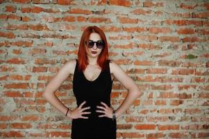 Red haired stylish girl in sunglasses wear in black, against abadoned place with brick walls. photo