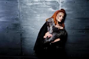 Fashion model red haired girl with originally make up like leopard predator wear on furs against steel wall. Studio portrait. photo