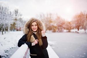 Beautiful brunette girl in winter warm clothing. Model on winter jacket against frozen lake at park.