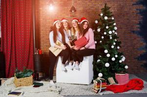 cuatro lindas amigas usan suéteres cálidos, pantalones negros y sombreros de santa contra el árbol de año nuevo con decoración navideña en el estudio. foto