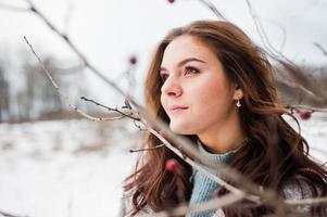 Close up portrait of gentle girl in gray coat near the branches of a snow-covered tree.