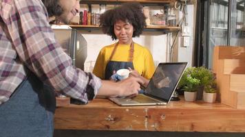 twee startende café-partners en vrienden, Afro-Amerikaanse vrouwen en Thaise mannelijke barista's praten en lachen vrolijk samen aan de balie van de coffeeshop, een vrolijke servicebaan en een MKB-ondernemer. video