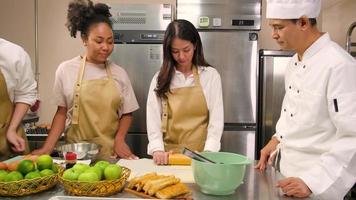 curso de culinária, chef masculino sênior em uniforme de cozinheiro ensina jovens estudantes de aula de culinária a amassar e enrolar massa de pastelaria, preparar ingredientes para alimentos de padaria, tortas de frutas na cozinha de aço inoxidável. video