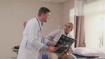 Caucasian male doctor in uniform health checks illness senior patient, diagnosis explains x-ray film in emergency room bed at hospital ward, elderly medical clinic, painful examination consultant.