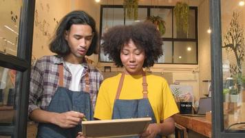 Two young startup barista partners with aprons stand at casual cafe door, write on board and show an open sign, happy and cheerful smiles with coffee shop service jobs, and new business entrepreneurs. video