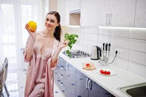 retrato de una mujer joven hermosa en túnica rosa posando con perejil y pimiento amarillo en sus manos en la cocina. foto