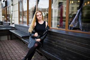 Blonde fashionable girl in long black leather coat sitting on bench. photo