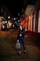 retrato nocturno de modelo de niña con gafas, jeans y chaqueta de cuero, con mochila, contra las luces de las calles de la ciudad. foto
