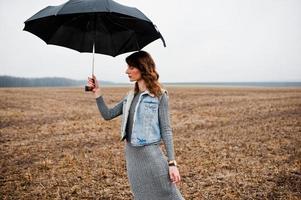 Portrait of brunette curly girl in jeans jacket with black umbrella at field. photo