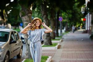 Portrait of a very attractive young woman in striped overall posing with her hat on a pavement in a town with trees in a background. photo