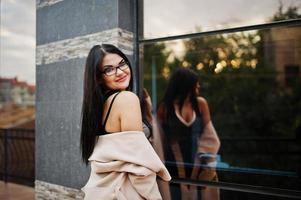 Black hair sexy woman in glasses and coat posed against building with modern windows. photo