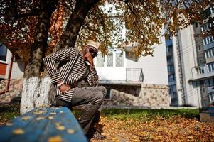 Portrait of stylish black african american man at hat and sunglasses against sunny autumn fall background. Rich people in africa at traditional dress. photo