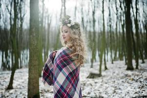 Curly cute blonde girl with wreath in checkered plaid at snowy forest in winter day. photo