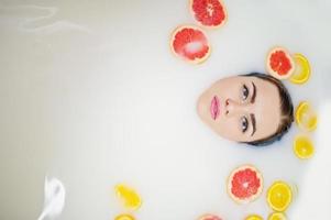 Sexy brunette girl at the bathroom with milk and fruits. Youth bath for body. photo