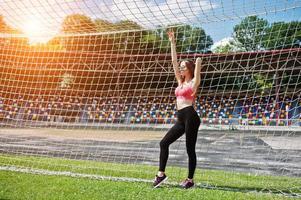 Fitness sporty girl in sportswear and sunglasses posed at stadium football gates outdoor sports. Happy sexy woman. photo