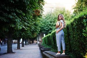 Portrait of a fabulous young woman in striped overall walking on the barrier in the park. photo