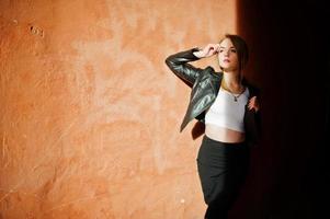 Elegant blonde girl wear on black leather jacket posing at streets of town background old wall with shadows. photo