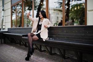 Black hair sexy woman in glasses and coat sitting on bench with mobile phone at hand and making selfie. photo