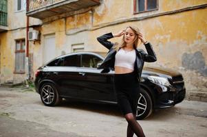 elegante chica rubia vestida con una chaqueta de cuero negro posando en las calles del coche de lujo de fondo de la ciudad. foto