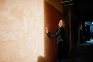 elegante chica rubia vestida con una chaqueta de cuero negro posando en las calles de la antigua muralla de fondo de la ciudad con sombras. foto