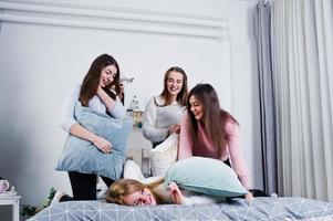 Four cute friends girls wear on warm sweaters and black pants on bed at new year decorated room on studio, play with pillows. photo