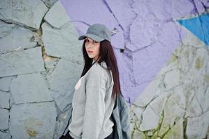 Stylish brunette girl in gray cap, casual street style on winter day against colored wall. photo