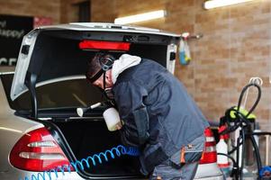 hombre con uniforme y respirador, trabajador del centro de lavado de autos, limpiando el interior del auto con un limpiador de vapor caliente. concepto de detalle de coche. foto