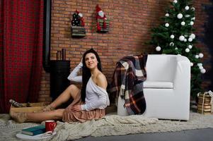 chica morena vestida posada cerca del árbol de año nuevo con decoración navideña en la sala de estudio de ladrillo. foto