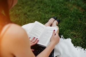 Close-up photo of woman's back while sitting on the blanket on the grass and reading a book.