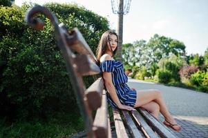 hermosa chica morena sentada en un banco en la calle de la ciudad vestida con un vestido azul a rayas. foto