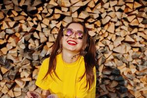 Young funny girl with bright make-up, tail hair wear on yellow shirt and sunglasses against wooden background. photo