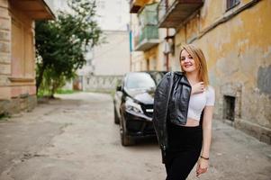 Elegant blonde girl wear on black leather jacket posing at streets of town background luxury car. photo