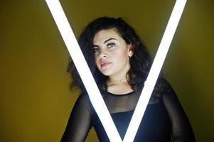 Curly brunette plus size model with two long led lamp against yellow wall. photo