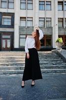 Portrait of a fabulous young successful woman in white blouse and broad black pants posing on the stairs with a huge white building on the background. photo