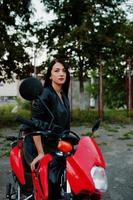 retrato de una mujer genial e impresionante con vestido y chaqueta de cuero negro sentada en una moto roja genial. foto