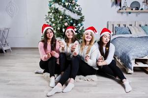 cuatro lindas amigas usan suéteres cálidos, pantalones negros y sombreros rojos de santa contra el árbol de año nuevo con decoración navideña en la habitación blanca y sostienen luces de bengala en las manos. foto