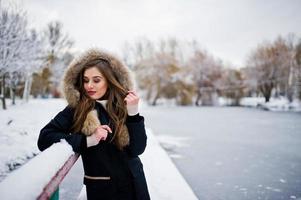 hermosa chica morena en ropa de abrigo de invierno. modelo en chaqueta de invierno contra el lago congelado en el parque. foto