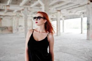 chica elegante de pelo rojo con gafas de sol vestidas de negro, contra un lugar abandonado. foto