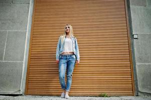 Blonde girl wear on jeans jacket with backpack posed against orange shutter. photo