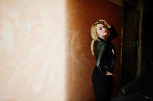 elegante chica rubia vestida con una chaqueta de cuero negro posando en las calles de la antigua muralla de fondo de la ciudad con sombras. foto