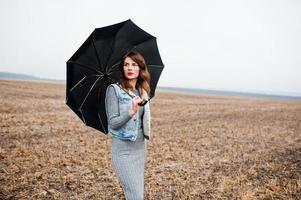 Portrait of brunette curly girl in jeans jacket with black umbrella at field. photo