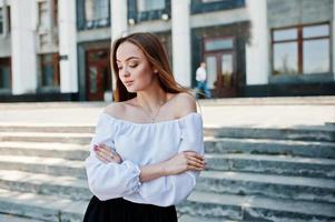 Portrait of a fabulous young successful woman in white blouse and broad black pants posing on the stairs with a huge white building on the background. photo