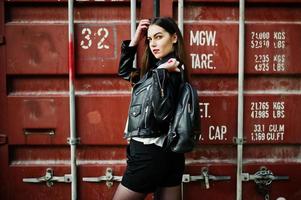 Stylish brunette girl wear on leather jacket and shorts with backpack against shipping container. photo