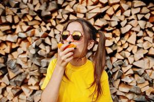 Young funny girl with bright make-up, tail hair wear on yellow shirt and sunglasses against wooden background eat orange.