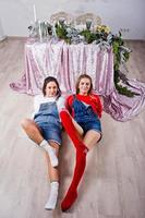 Two fun beautiful girls friends wear in overalls jeans shorts and gaiters against table with new year decoration. photo