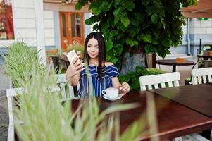 Gorgeous brunette girl sitting on the table in cafe with cup of coffee and making selfie. photo