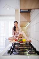 Happy young woman in pink robe sitting on the surface and making the salad. photo