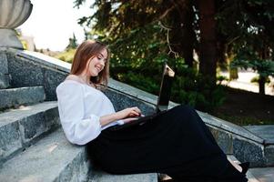 Good-looking young woman in white blouse, wide black pants and black classic high heels sitting on stairs and working on her laptop. photo